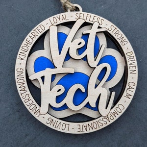 May include: Wooden ornament with a blue paw print and the words "Vet Tech" in a white circle. The circle is surrounded by a ring with the words "Kindhearted - Loyal - Selfless - Strong - Driven - Calm - Compassionate - Loving - Understanding".