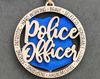 Puede incluir: Adorno de madera con una placa de policía azul y las palabras "Police Officer" en una fuente decorativa. El adorno está rodeado por un círculo de texto que dice "Patriótico, Fuerte, Inspirador, Digno de confianza, Leal, Honorable, Desinteresado, Valiente, Trabajador".
