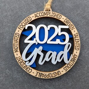 May include: A wooden ornament with a blue and silver graduation cap design. The ornament is inscribed with the year "2025" and the word "Grad". The ornament is also inscribed with the words "Motivated, Focused, Accomplished, Driven, Talented, Ambitious, Unstoppable, Confident".