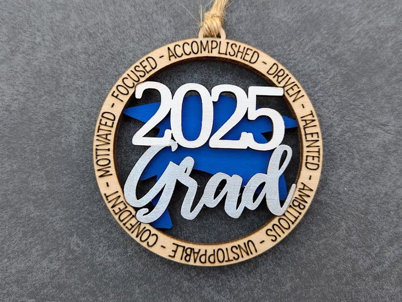 May include: A wooden ornament with a blue and silver graduation cap design. The ornament is inscribed with the year "2025" and the word "Grad". The ornament is also inscribed with the words "Motivated, Focused, Accomplished, Driven, Talented, Ambitious, Unstoppable, Confident".