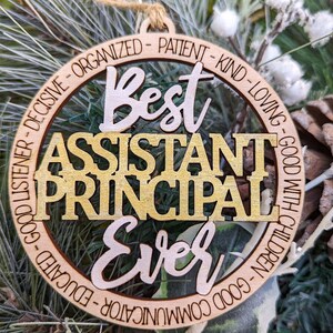 May include: A wooden ornament with the text "Best Assistant Principal Ever" in gold and brown lettering. The ornament is decorated with a circle of text that reads "Good Listener - Decisive - Organized - Patient - Kind - Loving - Good With Children - Good Communicator - Educated".