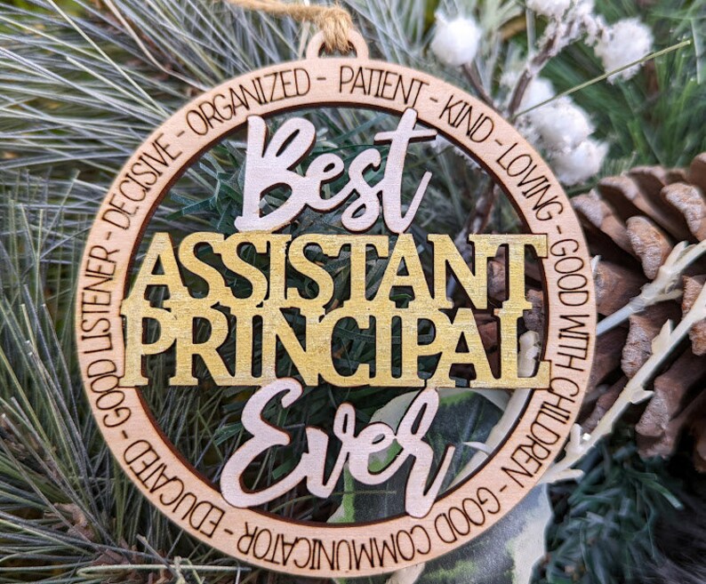 May include: A wooden ornament with the text "Best Assistant Principal Ever" in gold and brown lettering. The ornament is decorated with a circle of text that reads "Good Listener - Decisive - Organized - Patient - Kind - Loving - Good With Children - Good Communicator - Educated".