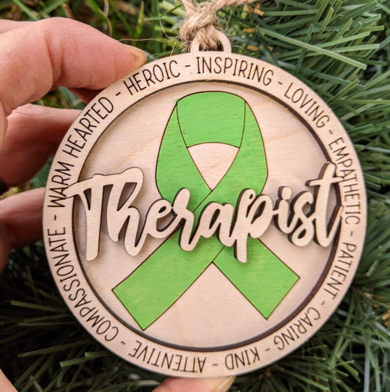 May include: Wooden ornament with a green ribbon and the word "Therapist" in a decorative font. The ornament is surrounded by a circle of text that reads "Warm Hearted - Heroic - Inspiring - Loving - Empathetic - Patient - Caring - Kind - Attentive - Compassionate".