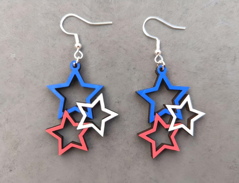May include: A pair of dangle earrings featuring three layered stars. The stars are red, white, and blue.