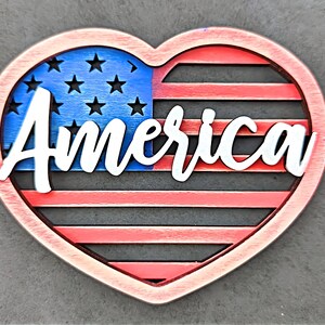 Może przedstawiać: Drewniany znak w kształcie serca z wzorem amerykańskiej flagi w kolorze czerwonym, białym i niebieskim. Słowo "America" jest napisane białymi literami kursywą na fladze.
