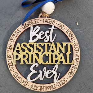 May include: A round wooden ornament with the text "Best Assistant Principal Ever" in gold and white lettering. The ornament is decorated with a black background and a ring of text around the outside.