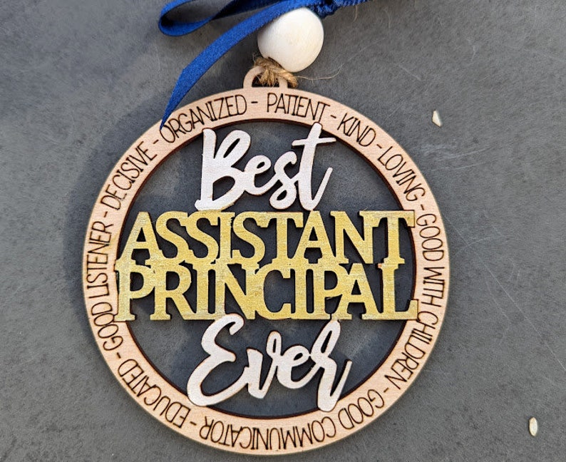 May include: A round wooden ornament with the text "Best Assistant Principal Ever" in gold and white lettering. The ornament is decorated with a black background and a ring of text around the outside.