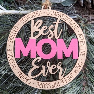 Puede incluir: Un adorno redondo de madera con las palabras "Best Mom Ever" en una fuente cursiva. La palabra "Mom" est&aacute; en rosa y el resto del texto est&aacute; en marr&oacute;n. El adorno est&aacute; rodeado por un c&iacute;rculo de texto que dice "Dedicado, Compasivo, Atento, Tranquilo bajo presi&oacute;n, Cuidadoso, Flexible, Habilidoso".