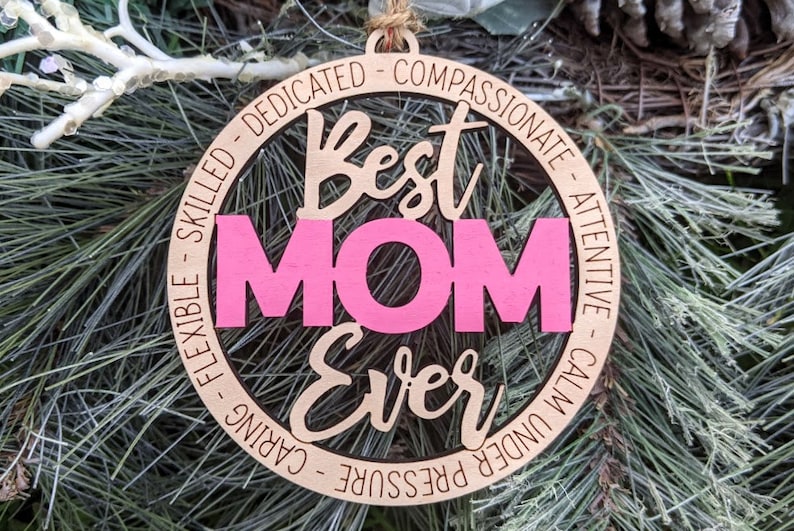 Puede incluir: Un adorno redondo de madera con las palabras "Best Mom Ever" en una fuente cursiva. La palabra "Mom" est&aacute; en rosa y el resto del texto est&aacute; en marr&oacute;n. El adorno est&aacute; rodeado por un c&iacute;rculo de texto que dice "Dedicado, Compasivo, Atento, Tranquilo bajo presi&oacute;n, Cuidadoso, Flexible, Habilidoso".