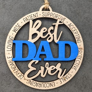 Puede incluir: Un adorno redondo de madera con las palabras "Best Dad Ever" en una fuente cursiva. La palabra "DAD" est&aacute; en azul y el resto del texto est&aacute; en marr&oacute;n. El adorno est&aacute; rodeado por un c&iacute;rculo de palabras que describen cualidades positivas, como "sabio", "paciente", "apoyo", "aceptando", "protector", "estable", "alentador", "fiable" y "amoroso".