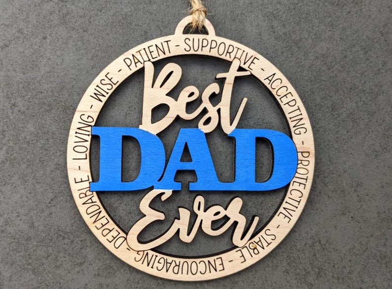 Puede incluir: Un adorno redondo de madera con las palabras "Best Dad Ever" en una fuente cursiva. La palabra "DAD" est&aacute; en azul y el resto del texto est&aacute; en marr&oacute;n. El adorno est&aacute; rodeado por un c&iacute;rculo de palabras que describen cualidades positivas, como "sabio", "paciente", "apoyo", "aceptando", "protector", "estable", "alentador", "fiable" y "amoroso".
