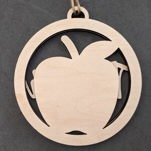 May include: A wooden ornament shaped like an apple with a cutout design. The ornament is a light brown color and has a string attached to the top for hanging.