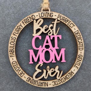 May include: A wooden circle ornament with the text "Best Cat Mom Ever" in pink and brown lettering. The circle is surrounded by the text "Fabulous Furry Friend - Loving - Purrfect - Dedicated - Gentle - Meowtastic - Nurturing - Devoted".