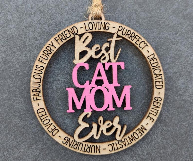 May include: A wooden circle ornament with the text "Best Cat Mom Ever" in pink and brown lettering. The circle is surrounded by the text "Fabulous Furry Friend - Loving - Purrfect - Dedicated - Gentle - Meowtastic - Nurturing - Devoted".