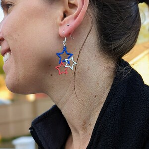 May include: A pair of dangle earrings featuring three layered stars. The stars are red, white, and blue.