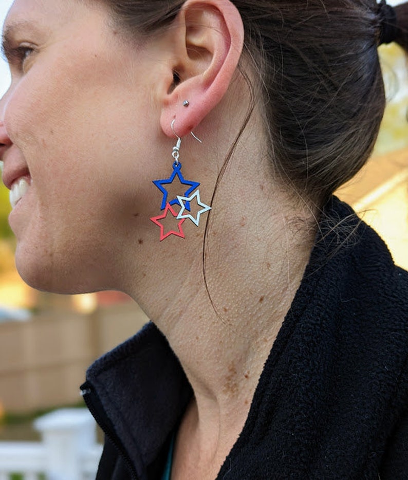 May include: A pair of dangle earrings featuring three layered stars. The stars are red, white, and blue.
