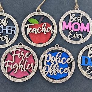 Puede incluir: Cinco adornos de madera con diferentes dise&ntilde;os y textos. Los adornos son redondos y tienen una cuerda para colgar. El texto en los adornos incluye "Mejor Maestro de Todos los Tiempos", "Mejor Mam&aacute; de Todos los Tiempos", "Mejor Pap&aacute; de Todos los Tiempos", "Bombero" y "Oficial de Polic&iacute;a".