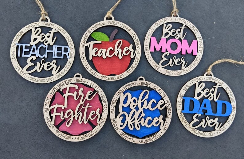 Puede incluir: Cinco adornos de madera con diferentes dise&ntilde;os y textos. Los adornos son redondos y tienen una cuerda para colgar. El texto en los adornos incluye "Mejor Maestro de Todos los Tiempos", "Mejor Mam&aacute; de Todos los Tiempos", "Mejor Pap&aacute; de Todos los Tiempos", "Bombero" y "Oficial de Polic&iacute;a".
