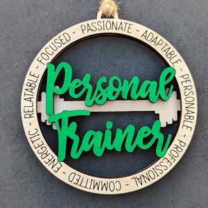 May include: Wooden ornament with the words "Personal Trainer" in green script, with a green and white dumbbell design. The ornament has a natural wood finish and a jute string for hanging. The outer ring has words like "focused" and "passionate".