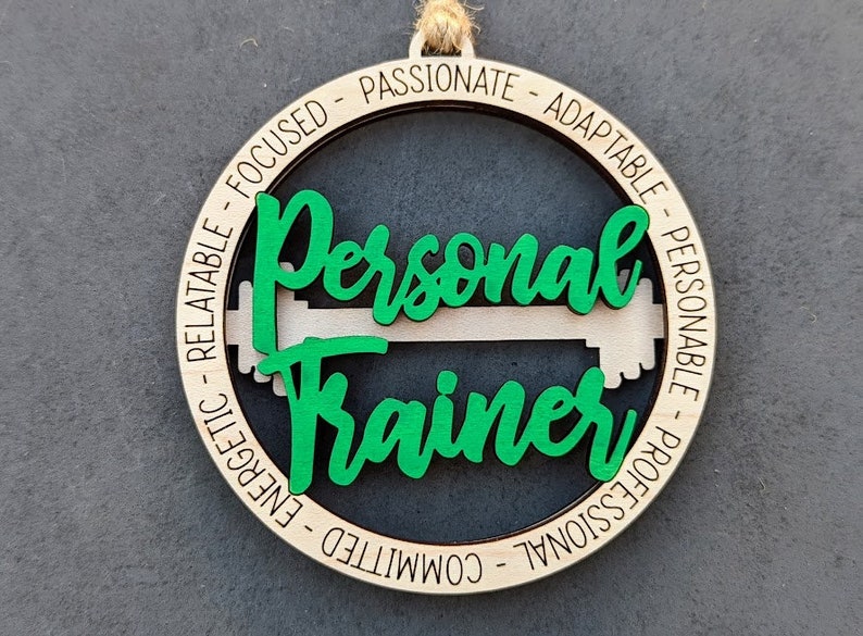 May include: Wooden ornament with the words "Personal Trainer" in green script, with a green and white dumbbell design. The ornament has a natural wood finish and a jute string for hanging. The outer ring has words like "focused" and "passionate".