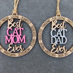 May include: Two wooden ornaments with a circle design. The top ornament says "Best Cat Mom Ever" in pink lettering. The bottom ornament says "Best Cat Dad Ever" in gray lettering.