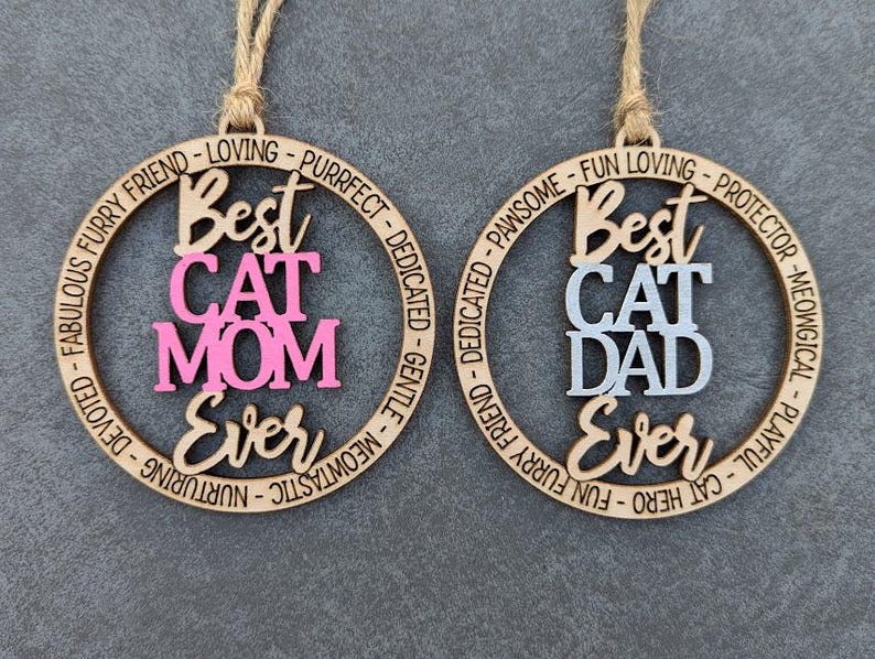 May include: Two wooden ornaments with a circle design. The top ornament says "Best Cat Mom Ever" in pink lettering. The bottom ornament says "Best Cat Dad Ever" in gray lettering.