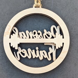 May include: A wooden ornament with the words "Personal Trainer" cut out in a circular design. The ornament is made of light-colored wood and has a natural twine loop for hanging. The design includes a barbell.