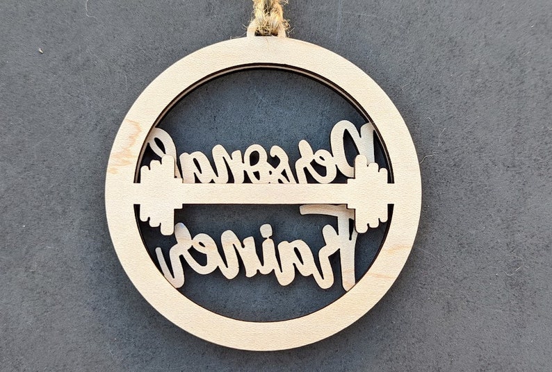 May include: A wooden ornament with the words "Personal Trainer" cut out in a circular design. The ornament is made of light-colored wood and has a natural twine loop for hanging. The design includes a barbell.