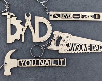 May include: Four wooden keychains with a father's day theme. The keychains are shaped like tools and have the words "Dad", "Level Headed", "Sawsome Dad", and "You Nail It".