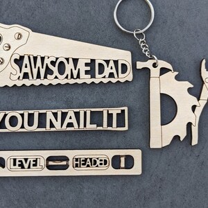 Puede incluir: Conjunto de cuatro llaveros de madera con formas de herramientas y juegos de palabras. Los llaveros dicen "Sawsome Dad", "You Nail It", "Level Headed" y "Dad" con una sierra, un martillo, un nivel y unas pinzas y un transportador.
