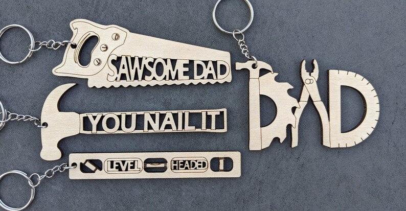 Puede incluir: Conjunto de cuatro llaveros de madera con formas de herramientas y juegos de palabras. Los llaveros dicen "Sawsome Dad", "You Nail It", "Level Headed" y "Dad" con una sierra, un martillo, un nivel y unas pinzas y un transportador.