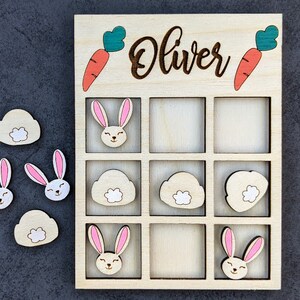 May include: Wooden game board with a grid of nine squares. The board is labeled "Oliver" and has images of carrots, bunnies, and clouds. There are also separate pieces with images of bunnies, carrots, and clouds.