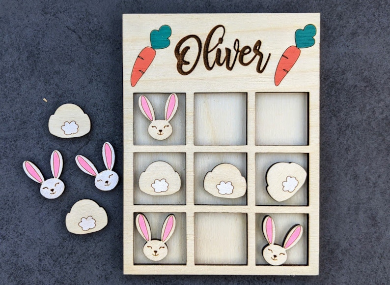 May include: Wooden game board with a grid of nine squares. The board is labeled "Oliver" and has images of carrots, bunnies, and clouds. There are also separate pieces with images of bunnies, carrots, and clouds.