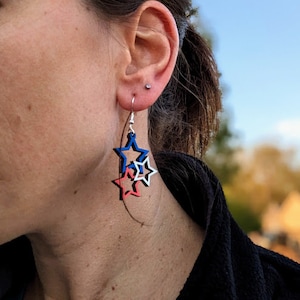 May include: A pair of dangle earrings featuring three layered stars. The stars are red, white, and blue.
