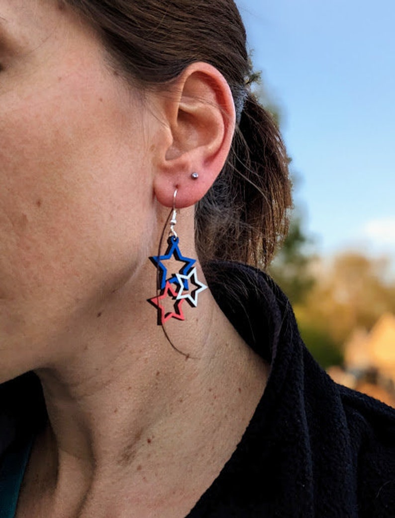 May include: A pair of dangle earrings featuring three layered stars. The stars are red, white, and blue.