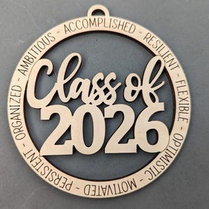 K&ouml;nnte beinhalten: Ein h&ouml;lzernes Abschlussornament mit der Aufschrift "Class of 2026" in einem kreisf&ouml;rmigen Design. Die W&ouml;rter "Organisiert - Ehrgeizig - Erfolgreich - Belastbar - Flexibel - Optimistisch - Motiviert - Beharrlich" sind eingraviert.