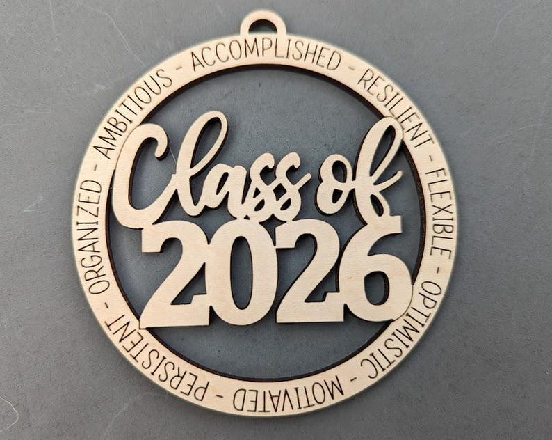K&ouml;nnte beinhalten: Ein h&ouml;lzernes Abschlussornament mit der Aufschrift "Class of 2026" in einem kreisf&ouml;rmigen Design. Die W&ouml;rter "Organisiert - Ehrgeizig - Erfolgreich - Belastbar - Flexibel - Optimistisch - Motiviert - Beharrlich" sind eingraviert.