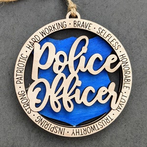 Puede incluir: Adorno de madera con una placa de polic&iacute;a azul en el centro y las palabras "Police Officer" en una fuente de escritura. El adorno est&aacute; rodeado por un c&iacute;rculo de texto que dice "Patri&oacute;tico, Trabajador, Valiente, Desinteresado, Honorable, Leal, Digno de confianza, Inspirador, Fuerte".