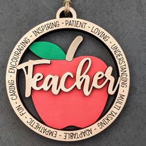 Puede incluir: Adorno de madera en forma de manzana con la palabra "Teacher" en el centro y una lista de adjetivos positivos alrededor del borde, incluyendo "adaptable", "emp&aacute;tico", "divertido", "cari&ntilde;oso", "alentador", "inspirador", "paciente", "amoroso", "comprensivo", "multitarea".