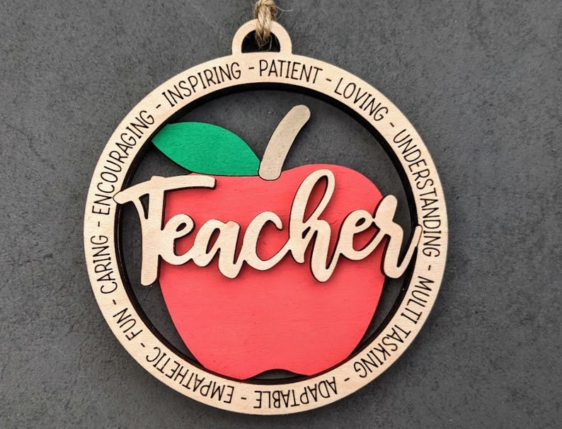 Puede incluir: Adorno de madera en forma de manzana con la palabra "Teacher" en el centro y una lista de adjetivos positivos alrededor del borde, incluyendo "adaptable", "emp&aacute;tico", "divertido", "cari&ntilde;oso", "alentador", "inspirador", "paciente", "amoroso", "comprensivo", "multitarea".