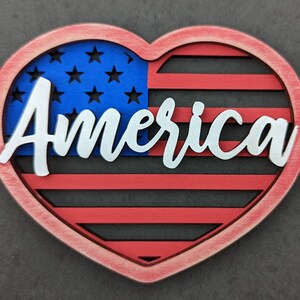 Może przedstawiać: Drewniany znak w kształcie serca w kolorze czerwonym, białym i niebieskim z napisem "America" białymi literami. Znak przedstawia stylizowany wz&oacute;r amerykańskiej flagi.
