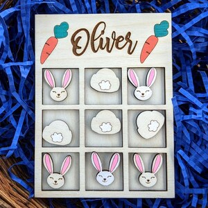 May include: Wooden game board with a tic-tac-toe grid. The board is painted white and has a brown border. The name "Oliver" is written in brown lettering at the top of the board. There are nine spaces on the board, each with a different image of a bunny or a cloud. The images are painted in shades of brown, pink, and white.