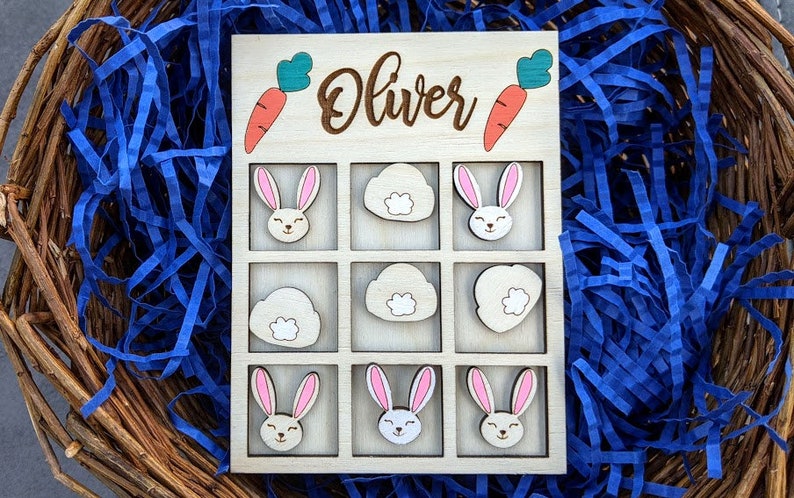 May include: Wooden game board with a tic-tac-toe grid. The board is painted white and has a brown border. The name "Oliver" is written in brown lettering at the top of the board. There are nine spaces on the board, each with a different image of a bunny or a cloud. The images are painted in shades of brown, pink, and white.