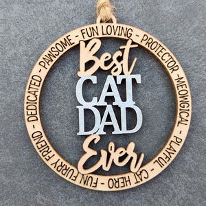 May include: A wooden circle ornament with the text "Best Cat Dad Ever" in white lettering. The circle is surrounded by a ring of text that reads "Dedicated - Pawsome - Fun Loving - Protector - Meowingical - Playful - Cat Hero - Fun Furry Friend."