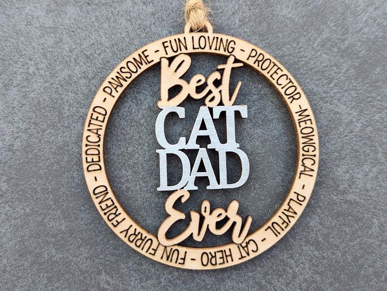 May include: A wooden circle ornament with the text "Best Cat Dad Ever" in white lettering. The circle is surrounded by a ring of text that reads "Dedicated - Pawsome - Fun Loving - Protector - Meowingical - Playful - Cat Hero - Fun Furry Friend."