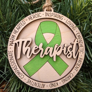 May include: A wooden ornament with a green ribbon in the center and the word "Therapist" written in a decorative font. The ornament is surrounded by a circle of text that reads: "Patient - Caring - Kind - Attentive - Compassionate - Warm Hearted - Heroic - Inspiring - Loving - Empathetic".
