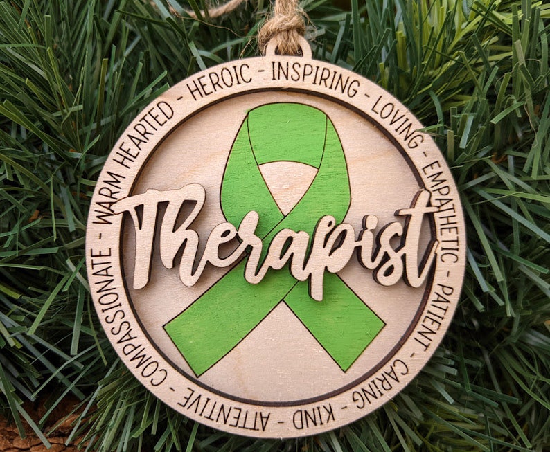May include: A wooden ornament with a green ribbon in the center and the word "Therapist" written in a decorative font. The ornament is surrounded by a circle of text that reads: "Patient - Caring - Kind - Attentive - Compassionate - Warm Hearted - Heroic - Inspiring - Loving - Empathetic".