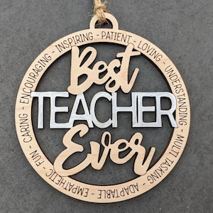Peut inclure: Une d&eacute;coration ronde en bois avec les mots "Best Teacher Ever" en argent et brun. La d&eacute;coration est entour&eacute;e d'un cercle de mots d&eacute;crivant des qualit&eacute;s positives d'enseignant, telles que "caring", "inspiring", "patient", "loving", "understanding", "multi-tasking", "adaptable", "empathetic", "fun", et "encouraging".