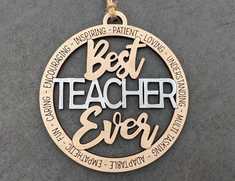 Peut inclure: Une d&eacute;coration ronde en bois avec les mots "Best Teacher Ever" en argent et brun. La d&eacute;coration est entour&eacute;e d'un cercle de mots d&eacute;crivant des qualit&eacute;s positives d'enseignant, telles que "caring", "inspiring", "patient", "loving", "understanding", "multi-tasking", "adaptable", "empathetic", "fun", et "encouraging".