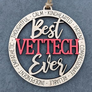 Könnte beinhalten: Ein runder Holzanhänger mit den Worten "Best Vet Tech Ever" in einer kursiv geschriebenen Schrift. Die Worte "Vet Tech" sind rot. Der Anhänger ist von einem Kreis aus Wörtern umgeben, die positive Eigenschaften beschreiben, wie "gedankenvoll", "ruhig", "gutherzig", "geduldig", "mitfühlend", "zuverlässig", "unabhängig" und "empathisch".